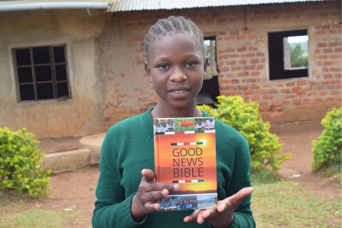 Keniaanse meisje staat voor een schoolgebouw met golfplaten en ramen zonder glas. Ze heeft een Bijbel in haar handen.